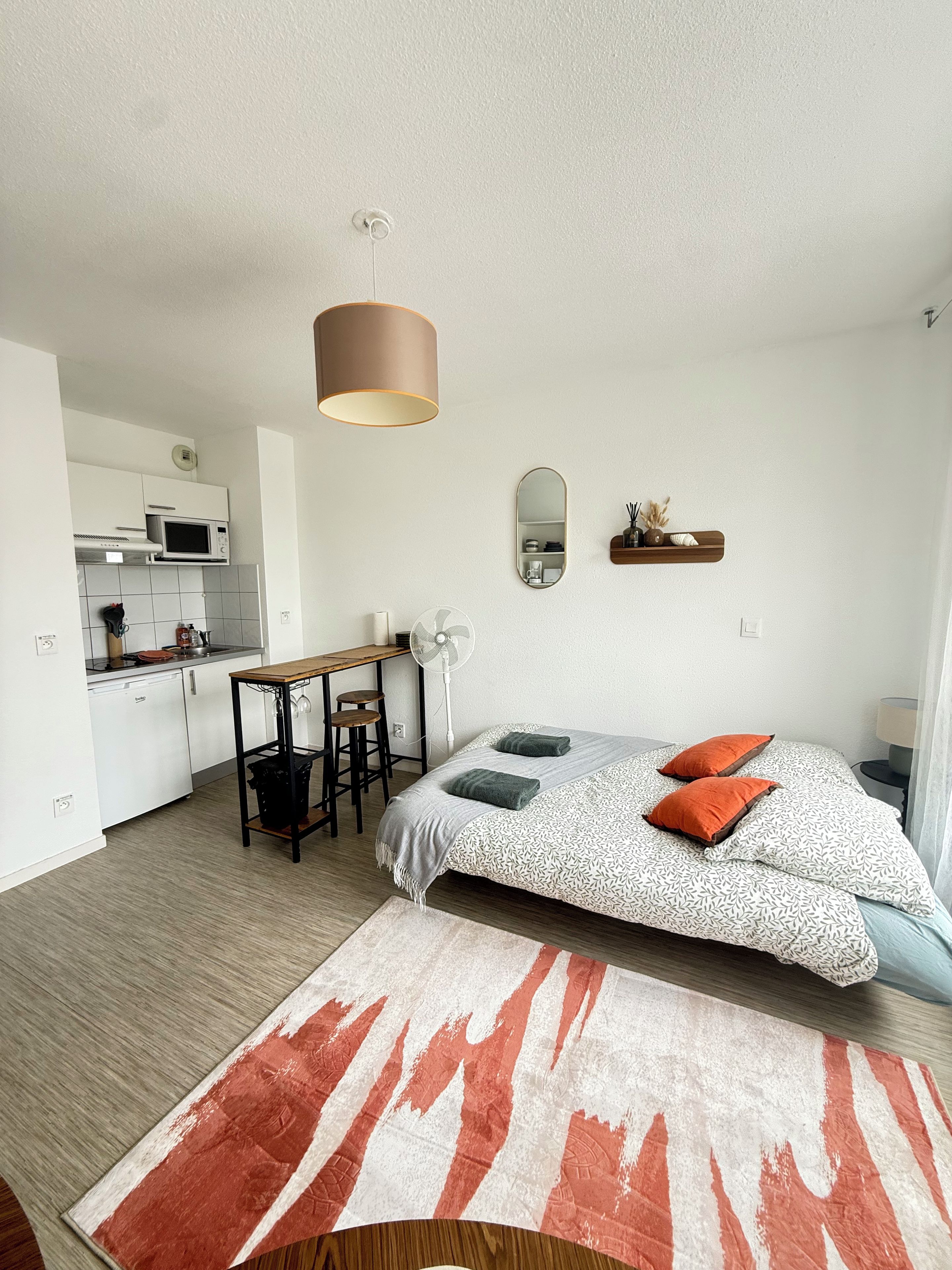 Une chambre d’hôtel compacte comprenant un coin cuisine, un lit avec une literie à motifs, une petite table et un miroir.