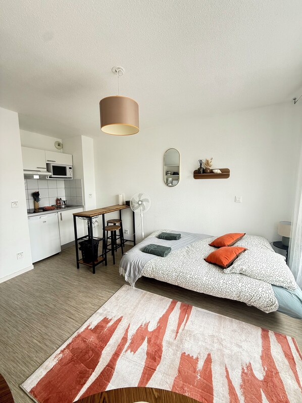 A compact hotel room with a kitchenette, a bed with patterned bedding, a small table, and a mirror.