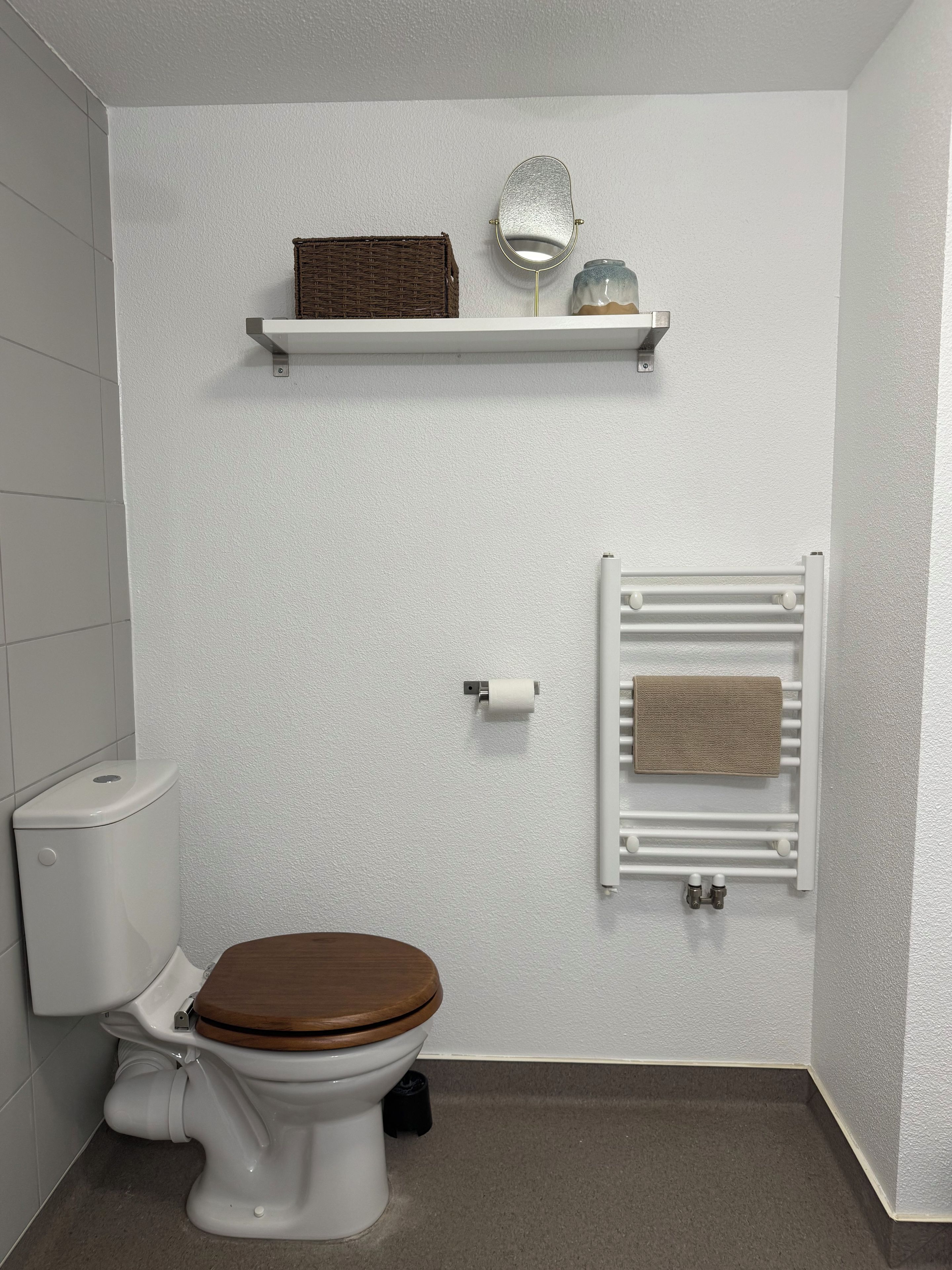 Une salle de bain avec des toilettes blanches, un abattant en bois, un porte-serviettes avec une serviette, une petite étagère avec un miroir et des objets décoratifs, et un porte-rouleau de papier toilette.