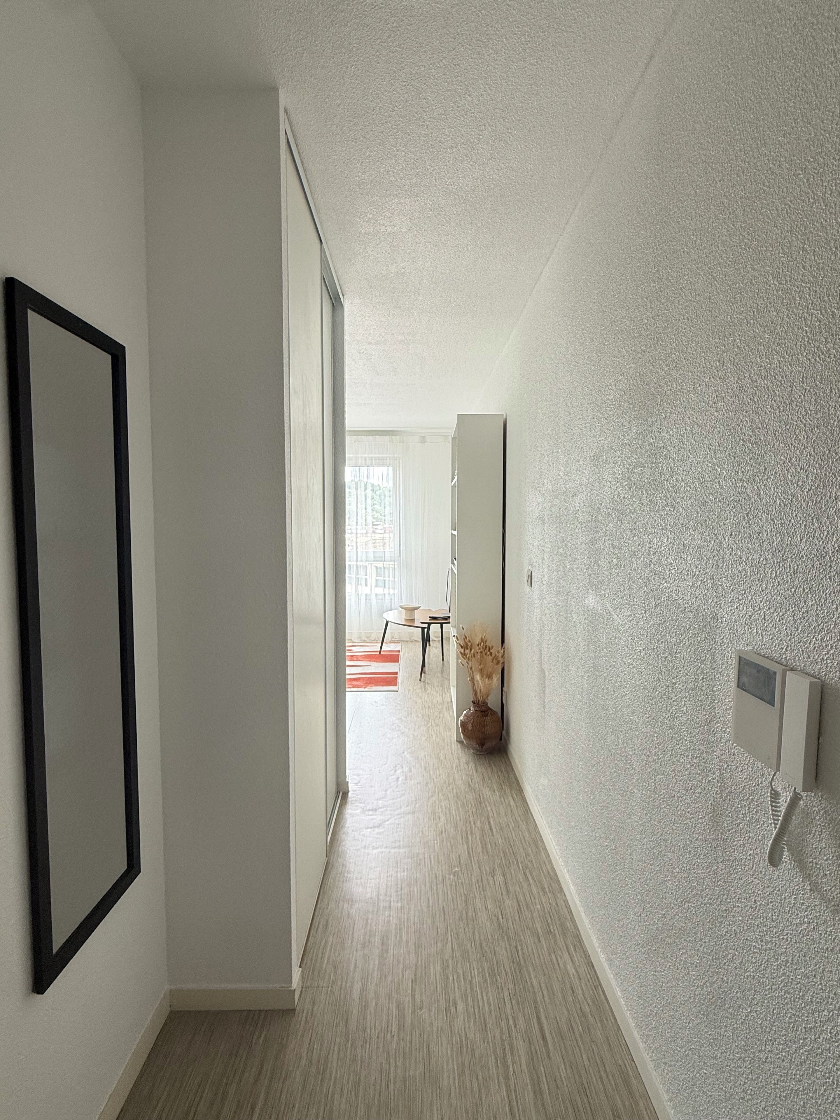 Un couloir doté d’un plancher en bois, avec un tableau encadré sur le côté gauche, un vase contenant des plantes séchées sur le côté droit, et une porte donnant sur une pièce où se trouvent une table et des chaises.