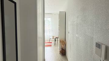 Un couloir doté d’un plancher en bois, avec un tableau encadré sur le côté gauche, un vase contenant des plantes séchées sur le côté droit, et une porte donnant sur une pièce où se trouvent une table et des chaises.