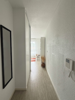 A hallway with a wooden floor, a framed picture on the left, a vase with dried plants on the right, and a door leading to a room with a table and chairs.