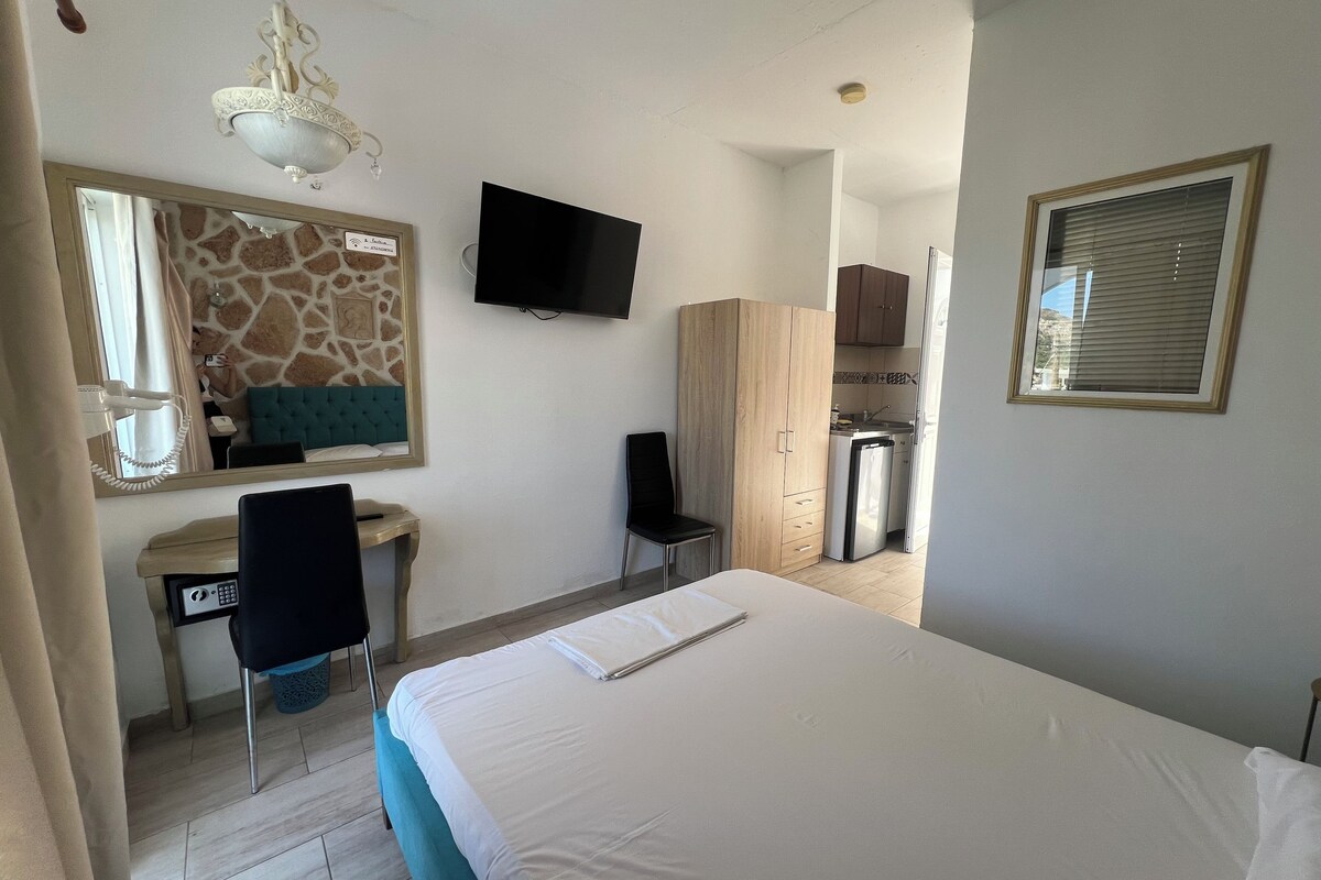 A hotel room with a bed, a desk with a chair, a television, a wardrobe, and a kitchenette.