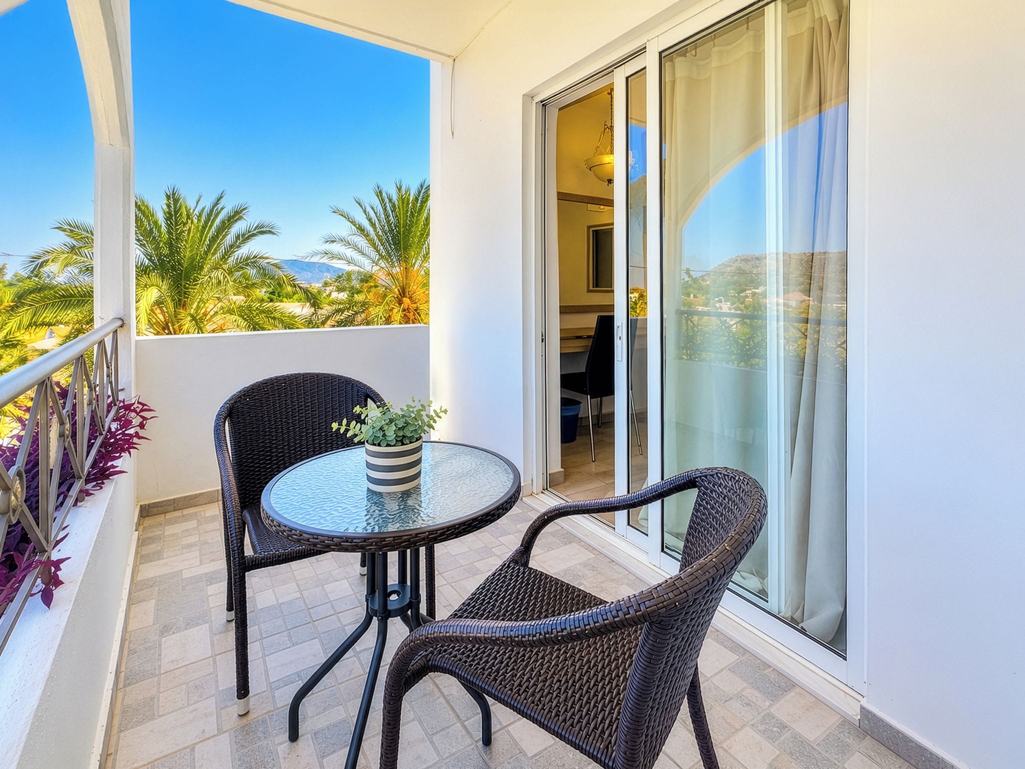 Un balcon avec une table en verre et deux fauteuils en osier, un pot de fleurs et une vue sur des palmiers et des montagnes.