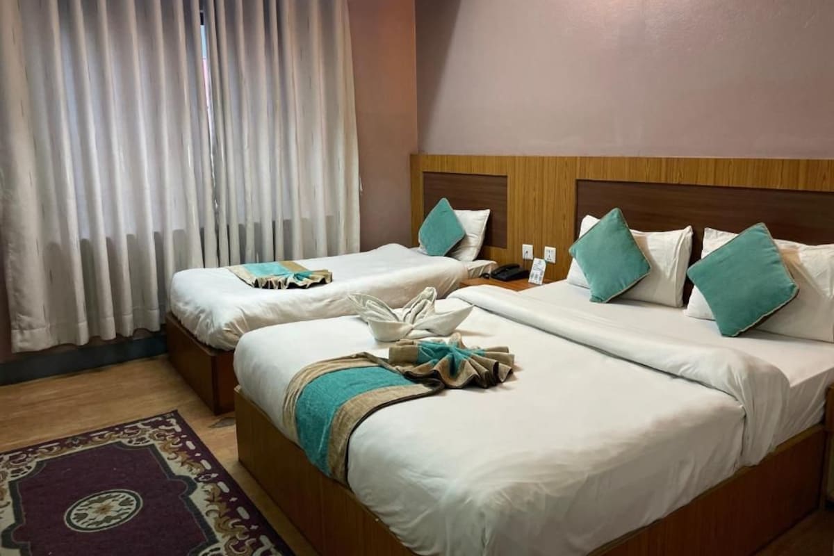 A hotel room with two beds, a wooden headboard, a window with white curtains, and a wall-mounted light fixture.