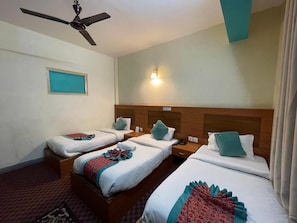 Habitación de hotel con tres camas individuales, ventilador de techo, mesita de noche e iluminación en la pared.