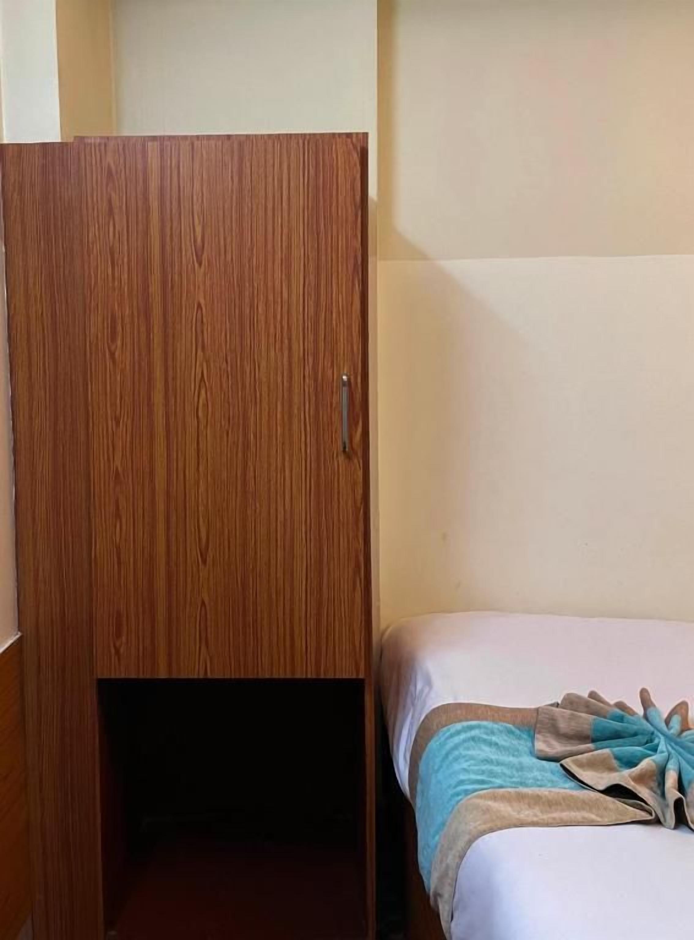 A room with a wooden wardrobe, a bed with a blue and beige blanket, and a white wall.