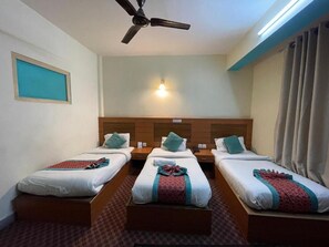 Habitación de hotel con tres camas individuales, un ventilador de techo, una lámpara fijada a la pared y un ventanal con marco azul.