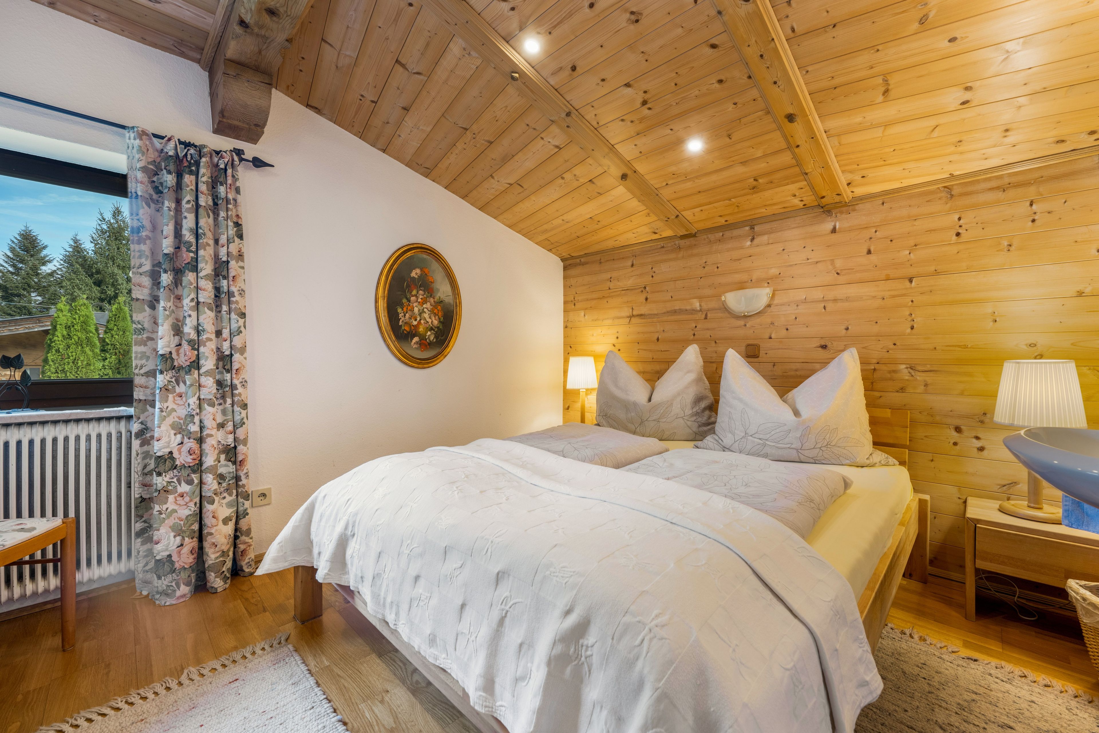 Ein Schlafzimmer mit Holzboden, einem Bett mit weißer Bettwäsche, einem Nachttisch mit Lampe und einem Fenster mit Vorhängen.