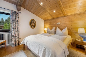 Ein Schlafzimmer mit Holzboden, einem Bett mit weißer Bettwäsche, einem Nachttisch mit Lampe und einem Fenster mit Vorhängen.