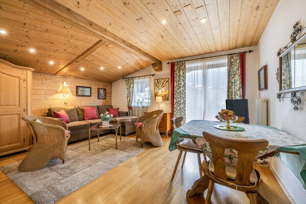 Ein gemütliches Interieur mit Holzbalkendecke, einer Essecke mit Tisch und Stühlen, einer Couch mit Kissen und einem Korbstuhl.