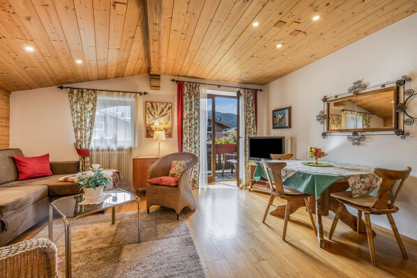 A cozy living room with a wooden ceiling, a sofa, a dining table with chairs, a television, and a window with curtains.