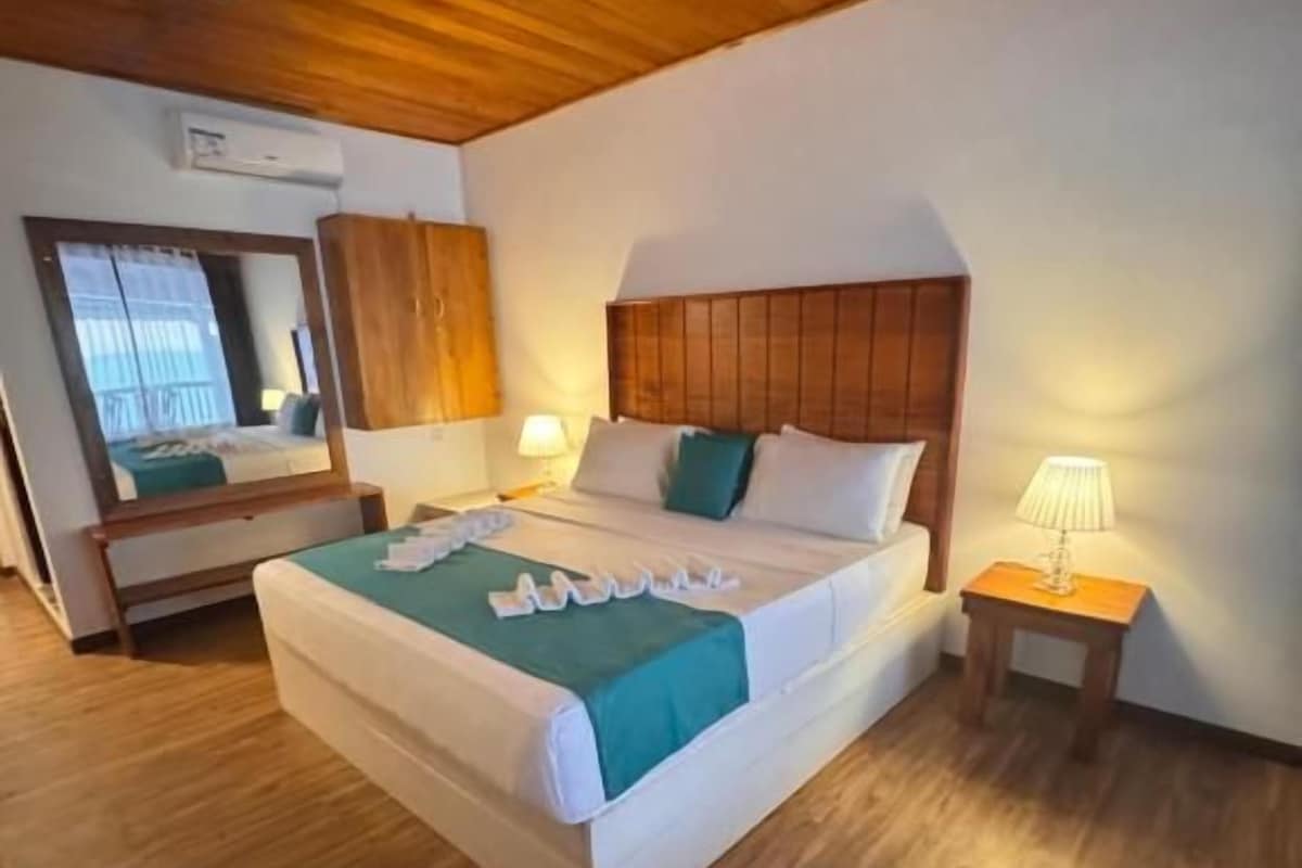Habitación de hotel con techo de madera, cama con cabecera blanca, mesita de noche con lámpara y espejo en la pared.