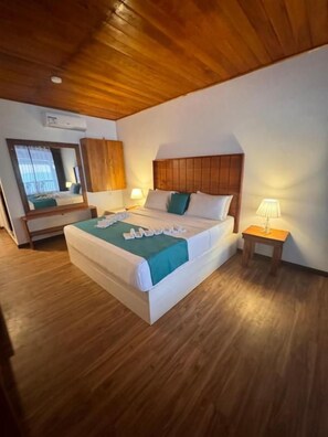Habitación de hotel con techo de madera, cama con cabecera blanca, mesita de noche con lámpara y espejo en la pared.