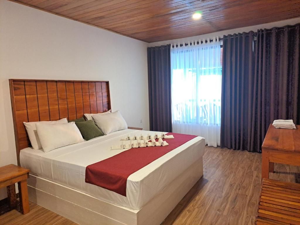 Una habitación de hotel con cabecera de madera, una cama con ropa blanca y una colcha roja, una mesita de noche de madera y una ventana con cortinas.
