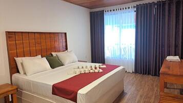 Una habitación de hotel con cabecera de madera, una cama con ropa blanca y una colcha roja, una mesita de noche de madera y una ventana con cortinas.