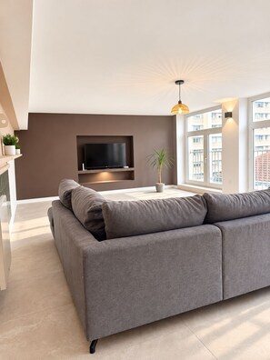 Un salon moderne avec un canapé gris, un téléviseur à écran plat fixé au mur et une grande fenêtre laissant entrer la lumière naturelle.