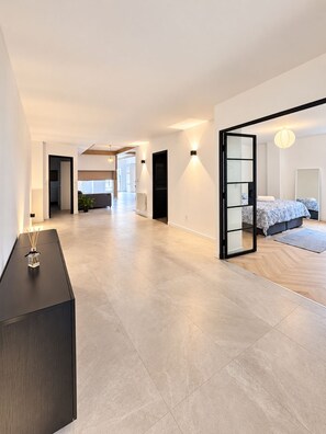 Un intérieur spacieux et moderne, doté d’un design minimaliste, comprenant un grand espace de vie ouvert, une chambre avec un lit et une table de chevet, ainsi qu’une console aux tons foncés agrémentée d’un objet décoratif.