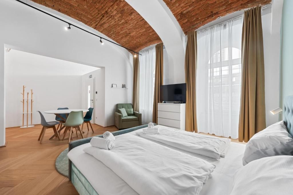 A modern hotel room with a brick ceiling, a flat-screen TV, a bed with white linens, a desk with chairs, and a dining area.