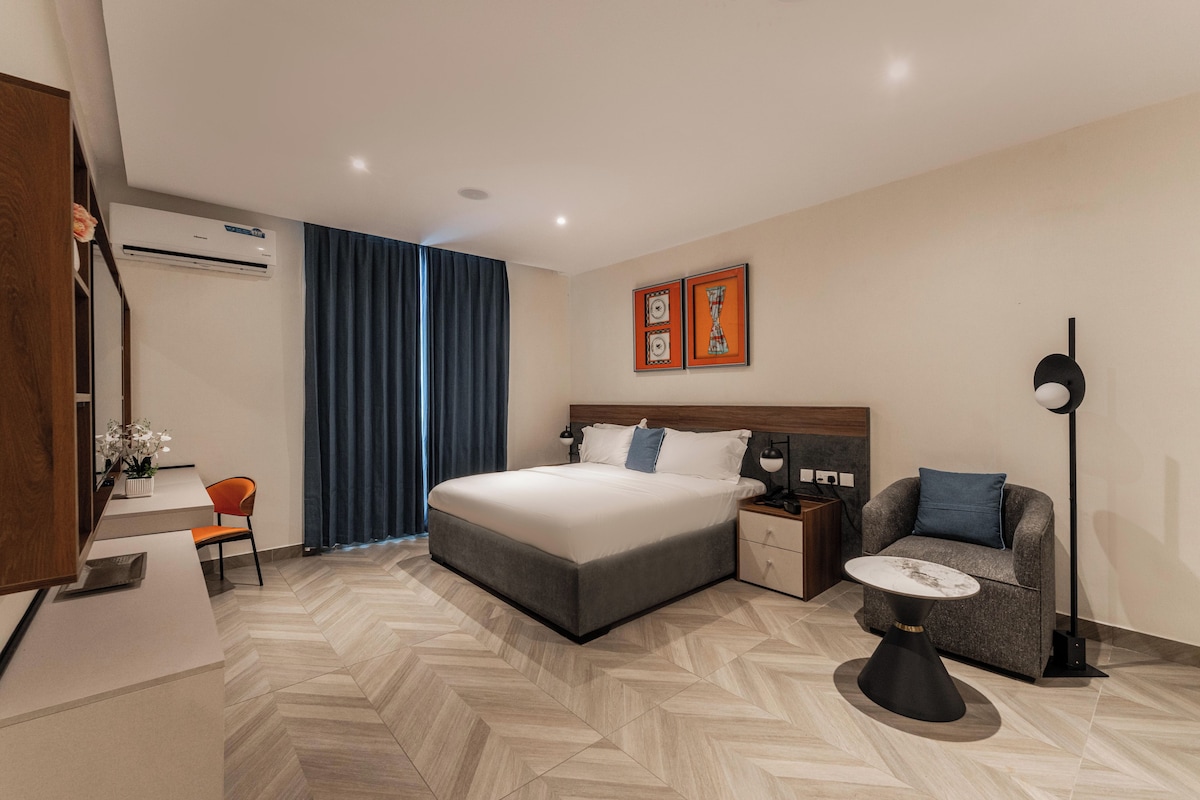Quarto de hotel moderno com cama, mesa de cabeceira, cadeira, escrivaninha e uma janela grande com cortinas.