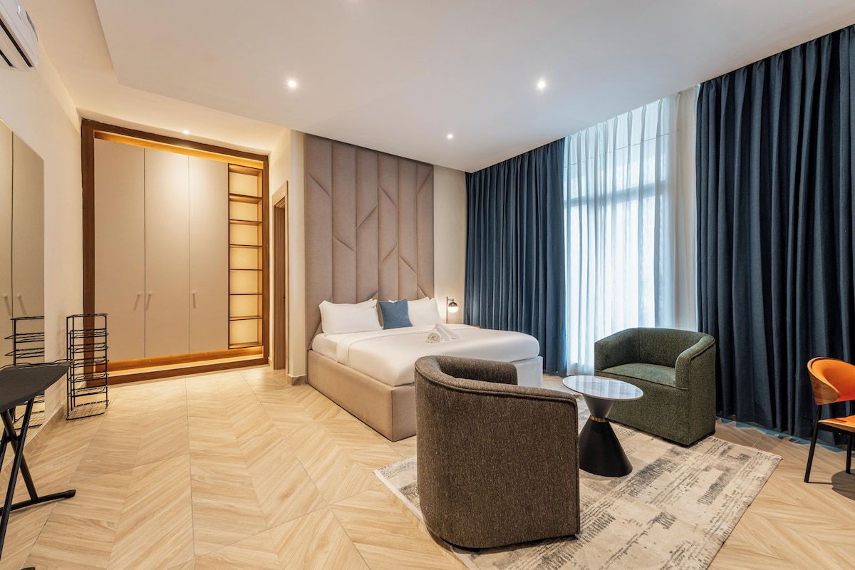 Quarto de hotel moderno com cama, área de estar com mesa e uma grande janela com cortinas.