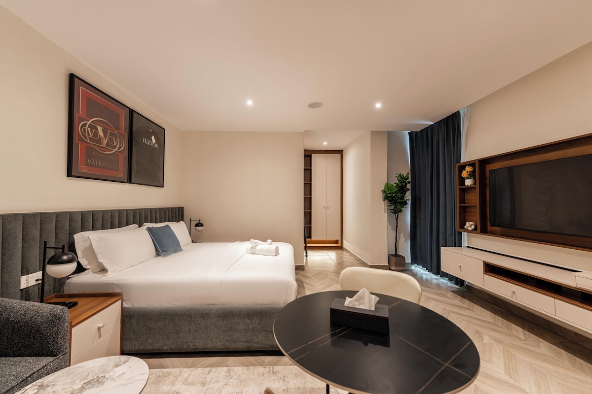 Quarto de hotel moderno com cama, mesa de cabeceira, luminária, TV e um espaço de estar com mesa e cadeiras.
