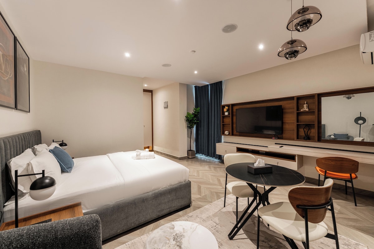 Quarto de hotel moderno com uma cama, escrivaninha, cadeira, televisão e espelho.