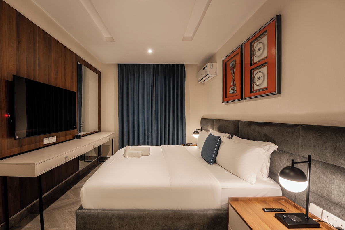 Quarto de hotel com uma cama grande, televisão de tela plana, escrivaninha com abajur e obras de arte nas paredes.