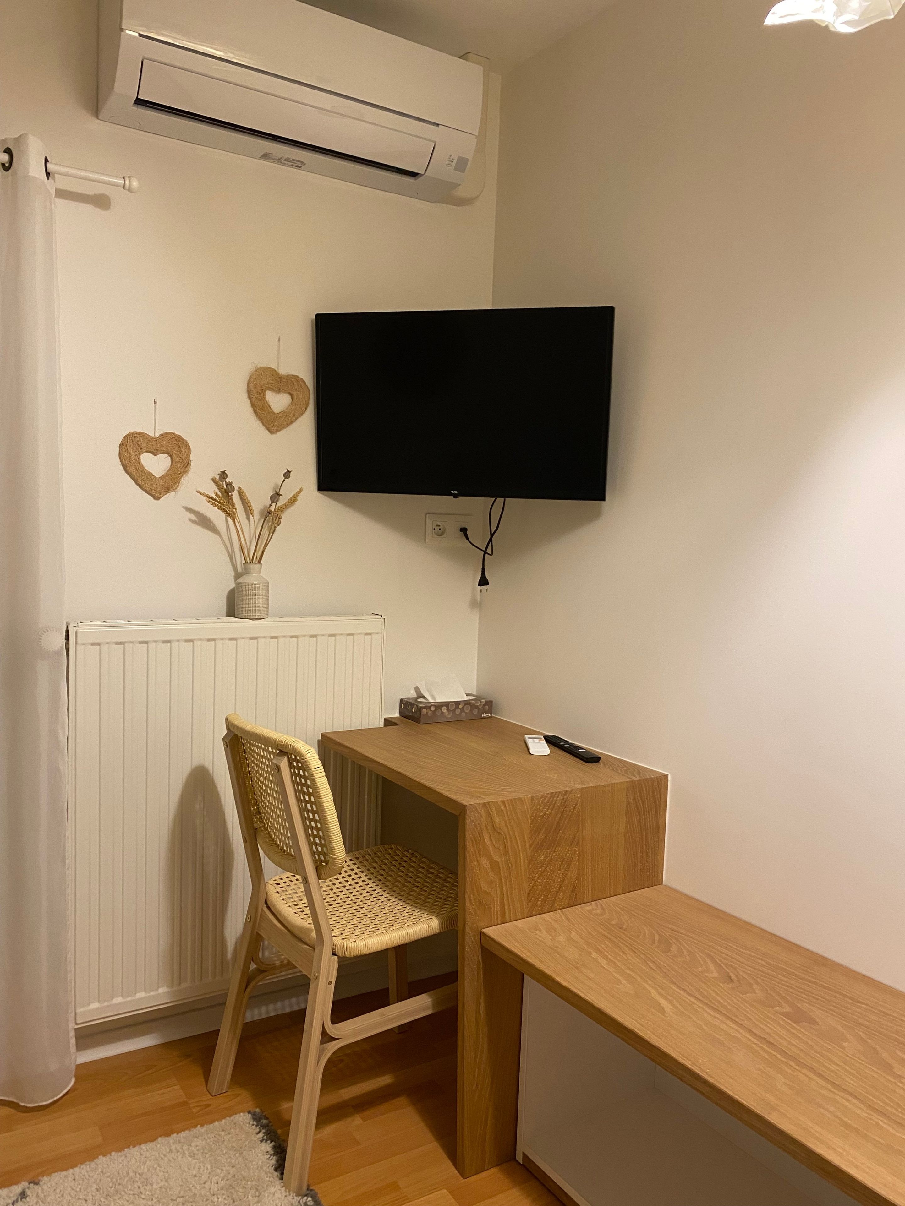 Une pièce avec un radiateur blanc, un téléviseur fixé au mur, un bureau en bois, une chaise en osier et un rideau blanc.