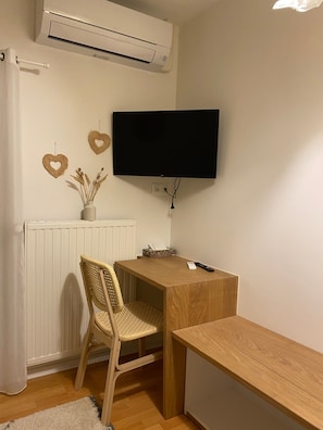 Habitació amb un radiador blanc, televisió penjada a la paret, escriptori de fusta, cadira de vimet i cortina blanca.