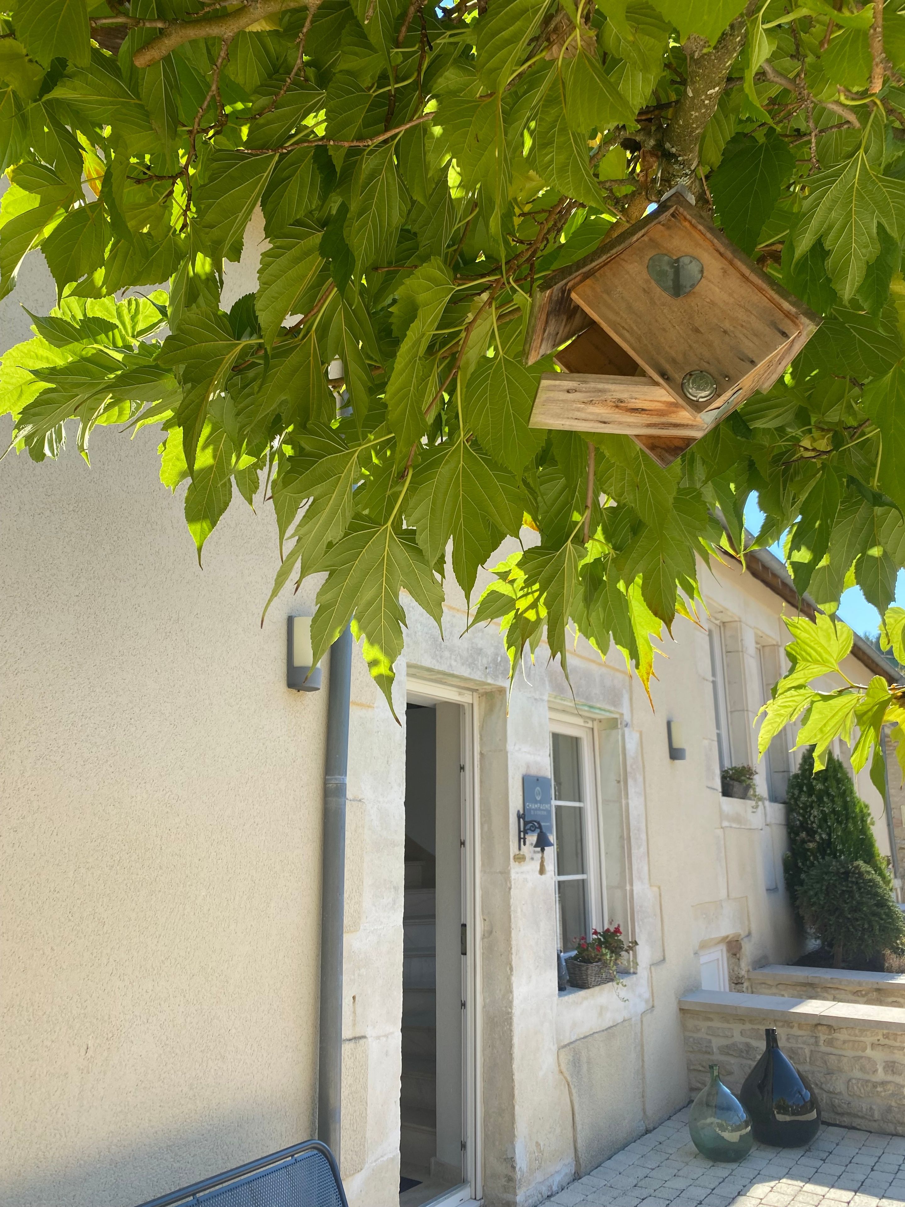 Un bâtiment à la façade en pierre, une mangeoire à oiseaux en bois suspendue à un arbre et une porte donnant sur un espace intérieur.