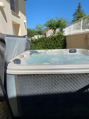 Un bain à remous extérieur doté d’un espace détente couvert, entouré d’une haie et d’une clôture blanche.