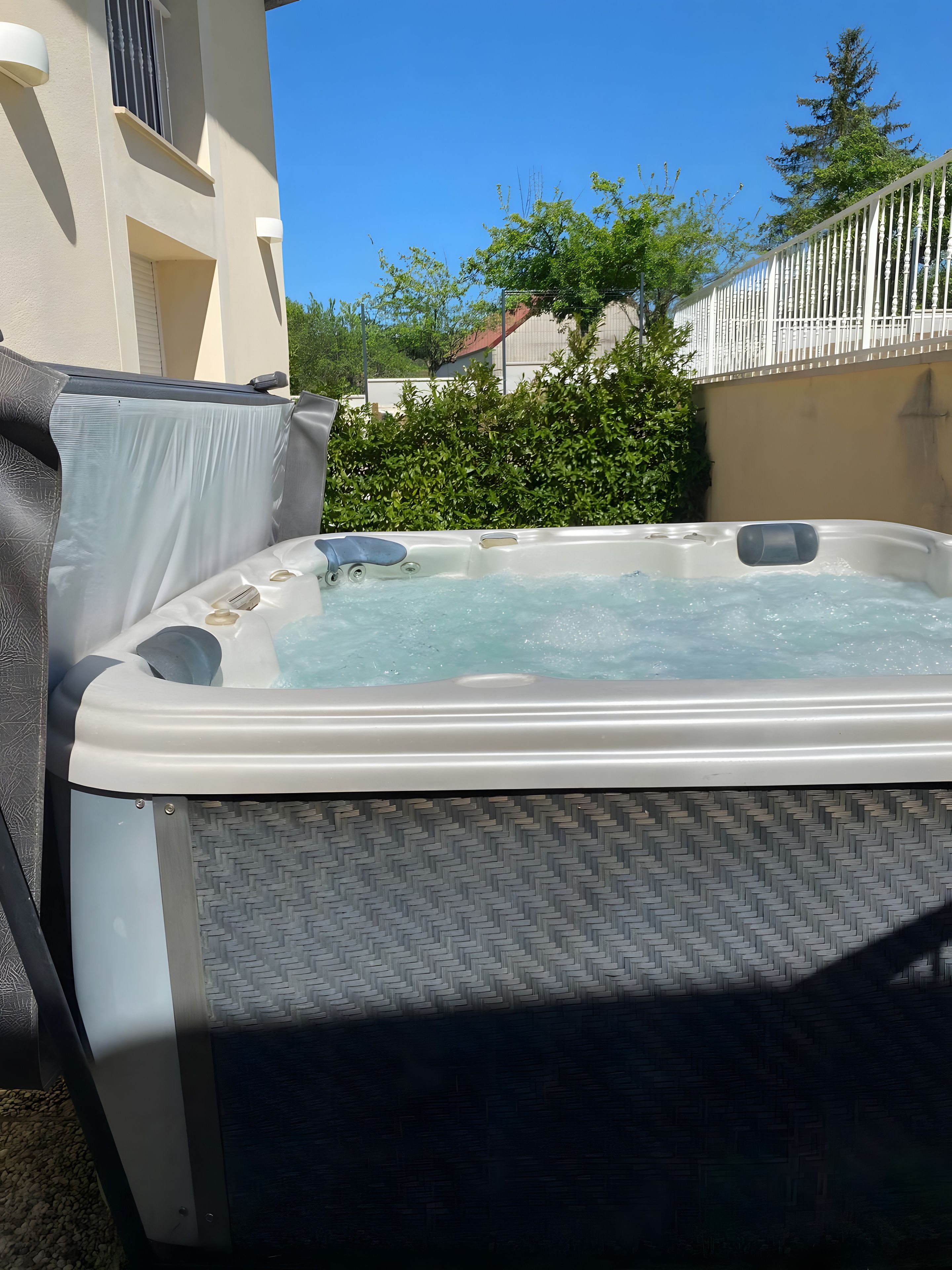 Un bain à remous extérieur doté d’un espace détente couvert, entouré d’une haie et d’une clôture blanche.