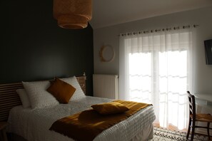 Une chambre à coucher avec un lit, une tête de lit en bois, un radiateur blanc, une fenêtre avec des rideaux légers et un téléviseur fixé au mur.