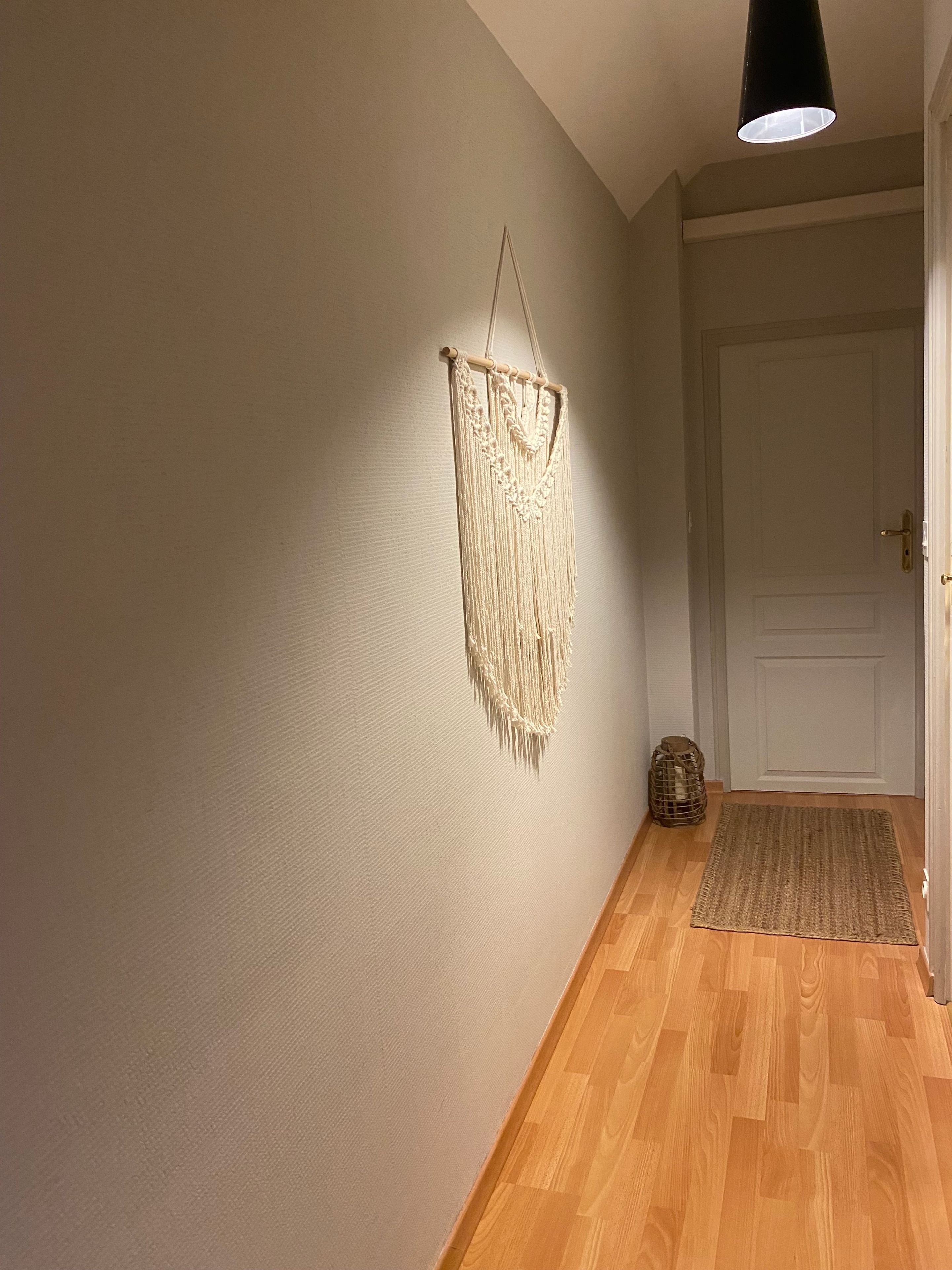 Un couloir orné d’une décoration murale en macramé, doté d’un plancher en bois, d’une porte et d’un petit panier.