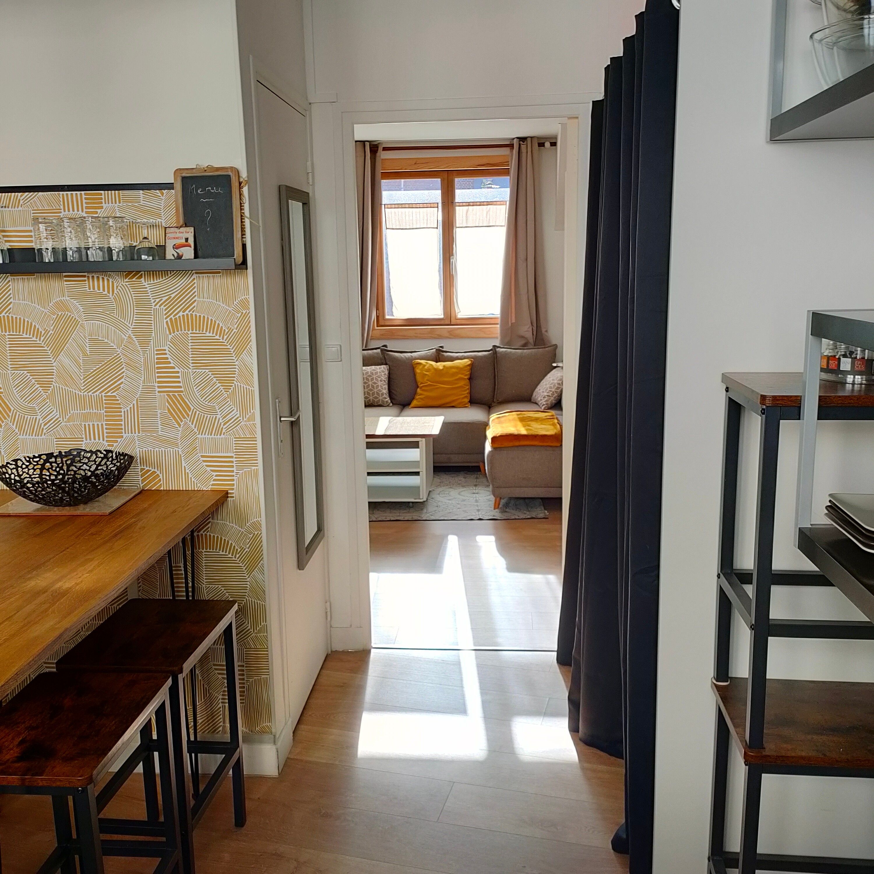 Ein schmaler Flur führt zu einem Wohnbereich mit einer Couch und Kissen, einem Essbereich mit Tisch und Stühlen sowie einer Küche mit einer Ablage.