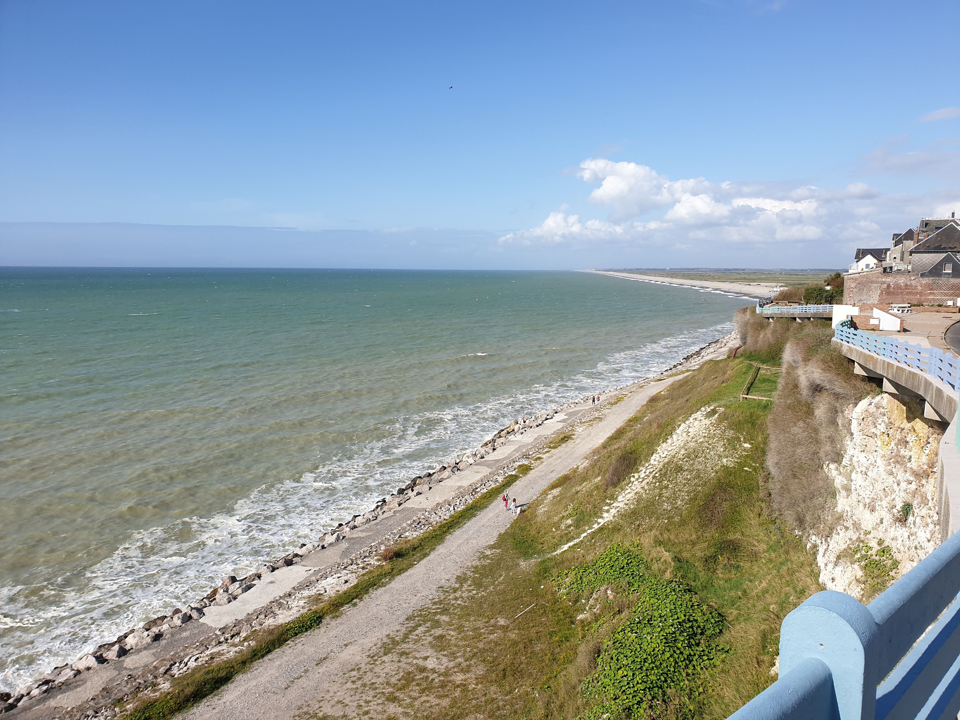 Udsigt over kysten med en strand, en sti og en bygning med gelænder i blå.