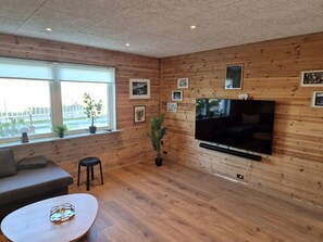 A modern living room with a large flat-screen TV mounted on the wall, wooden paneling, a sofa, a coffee table, and a window with a view of the city. - Hornagøta 4, 2 (Mid & Attic floor) (Thorshavn)
