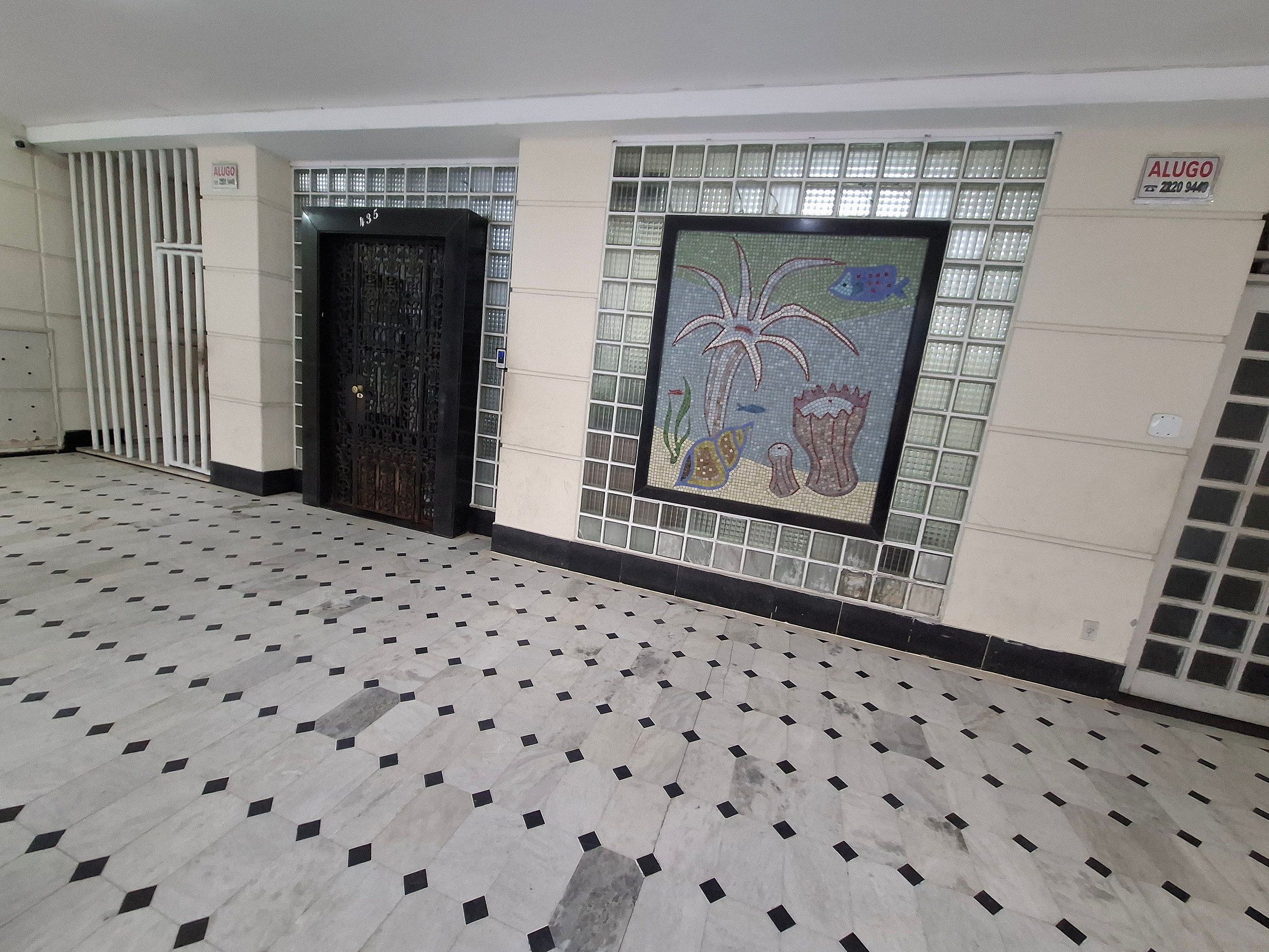 A hallway with a tiled floor, a decorative wall mosaic, a black door, and a sign with the name 'ALUGO'.