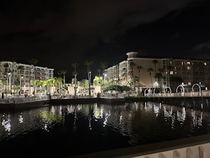 Vista nocturna de una zona costera con edificios, palmeras y una superficie acuática reflectante.