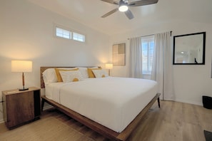 Quarto com cama, duas mesas de cabeceira, ventilador de teto, janela com cortinas e espelho fixado na parede.