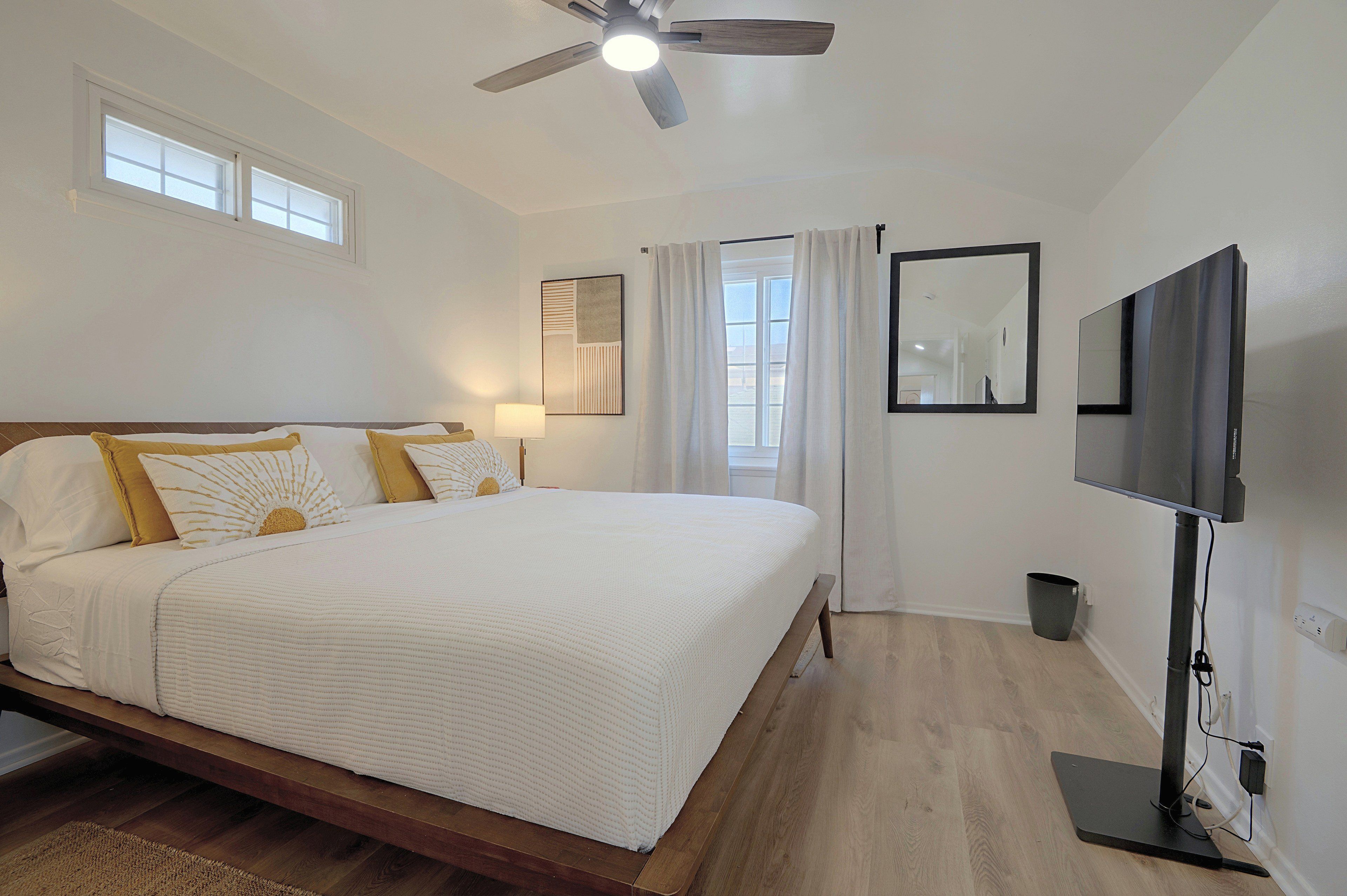A bedroom with a bed, a ceiling fan, a TV, and a window with curtains.