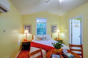 A bedroom with a bed, bedside lamps, a window with a view of trees, a door with a coat rack, and a small table with a plant and a card.