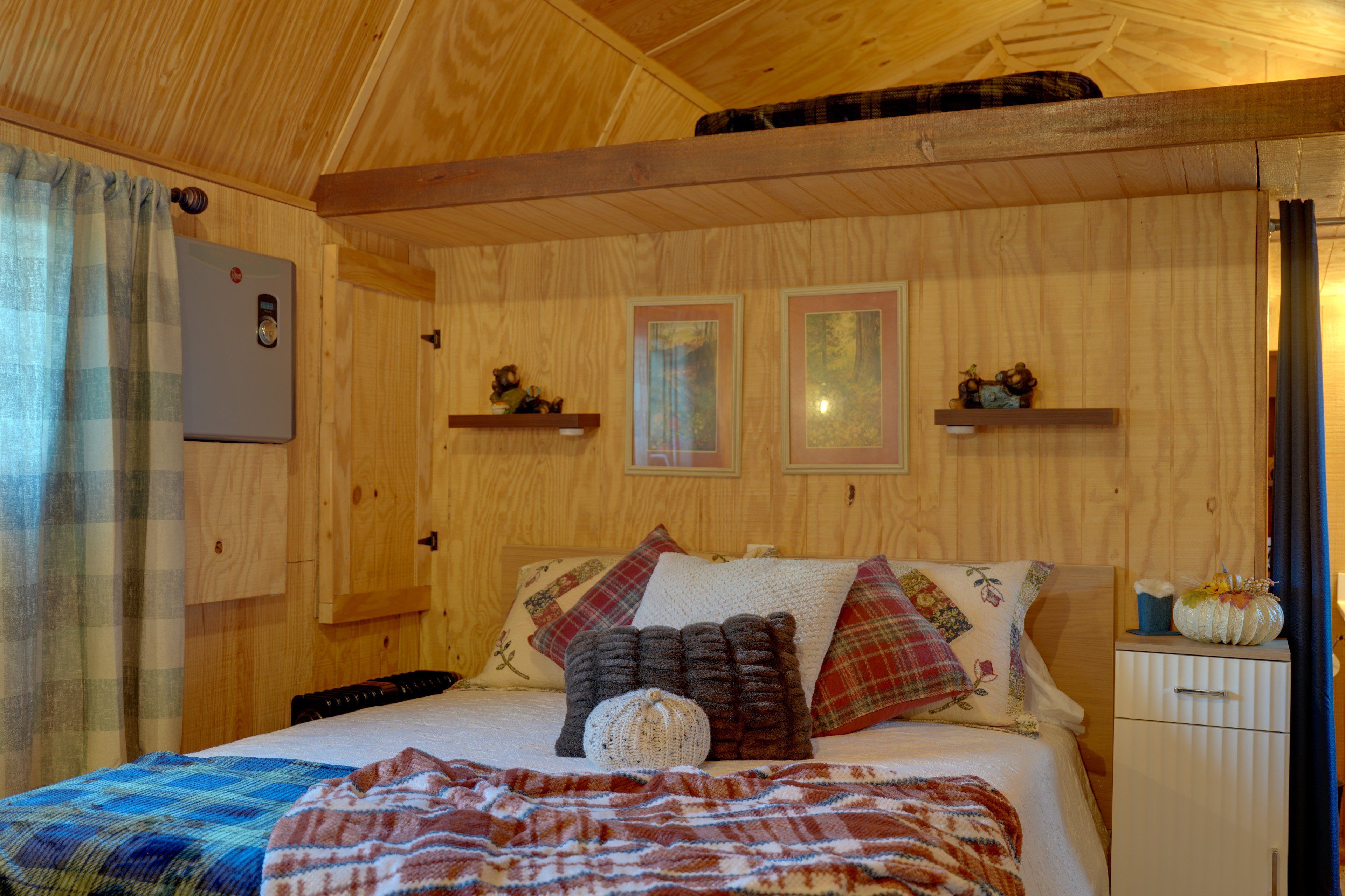 Ein Holzinterieur mit einem Bett, Kissen, einer Decke, einem Thermostat und gerahmten Bildern an der Wand.