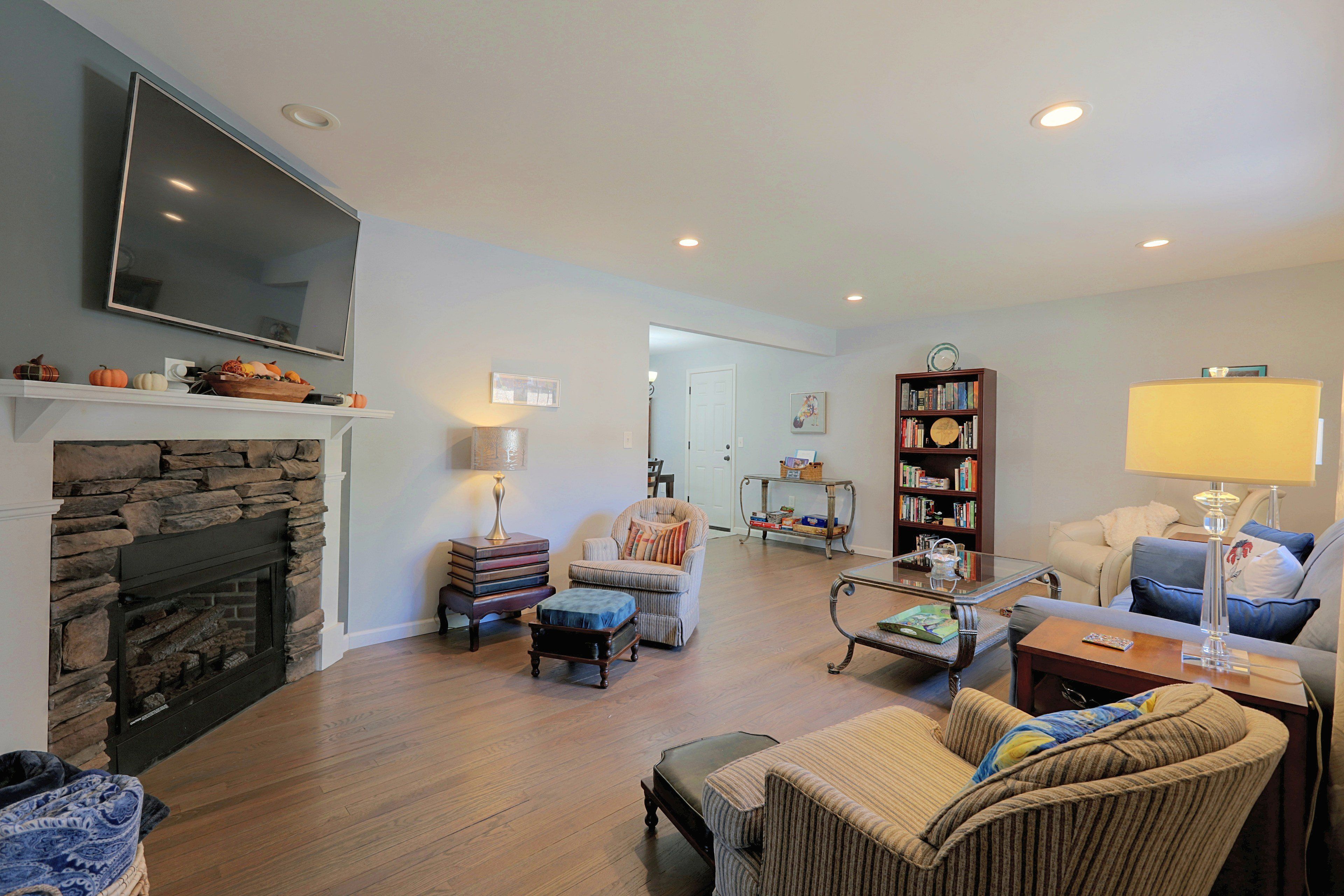 A living room with a stone fireplace, a television mounted on the wall, a bookshelf, and a comfortable seating area with a sofa and armchairs.
