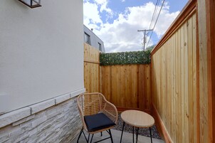 Un pequeño espacio al aire libre con una silla de mimbre y una mesa redonda, junto a una valla de madera y un edificio.