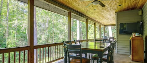 A covered outdoor deck with a dining table and chairs, surrounded by wooden railings, overlooking a forested area.