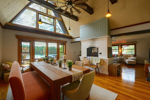 A spacious living room with a wooden dining table, comfortable seating, a fireplace, and large windows offering a view of the outdoors.