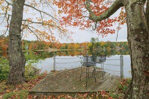 A wooden dock with a metal chair, surrounded by trees with autumn leaves, overlooking a calm lake.