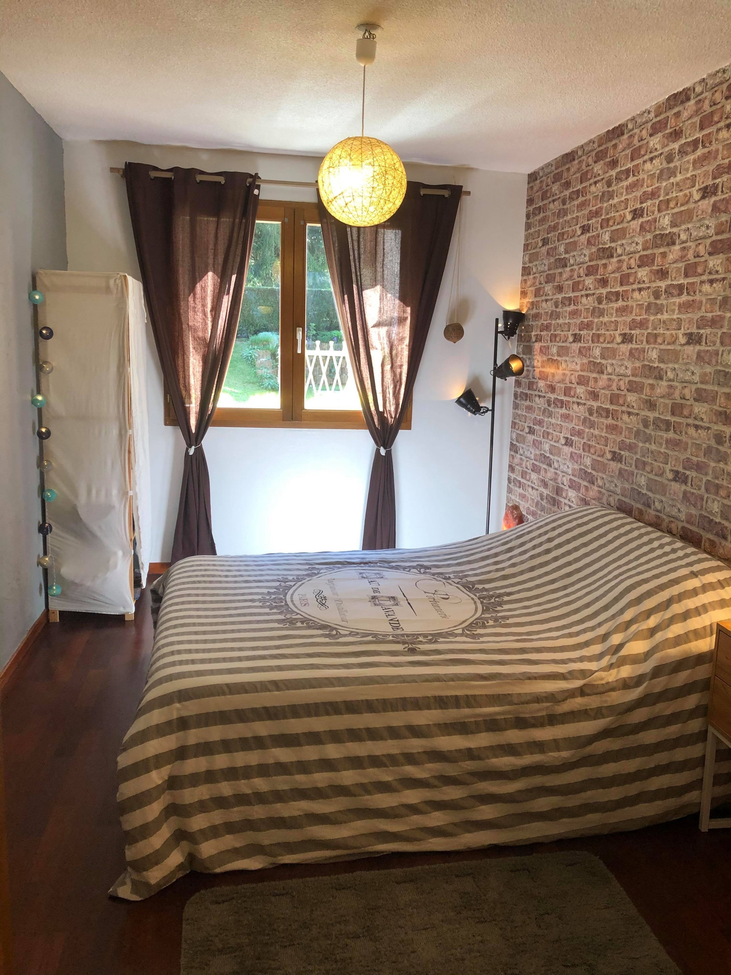 A bedroom with a bed, curtains, a hanging light fixture, a brick wall, and a window.